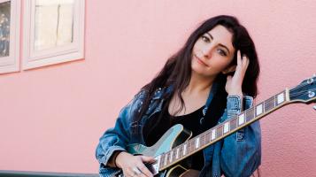 AliT stands in front of a pink wall holding an electric guitar.