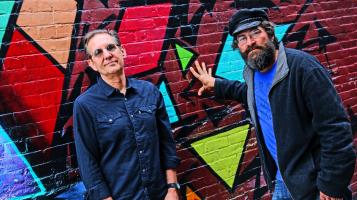 The two members of Soule Monde stand in front of a colorful mural of abstract shapes.