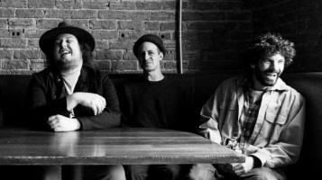 Troy Milette and his two bandmates sit at a table in front of a brick wall.