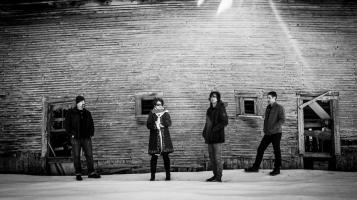 Shown in black and white, the four members of Maple Run stand in front of a dilapidated barn wall.