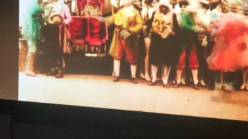 A screen displaying the a distorted image of a group of people in brightly colored eighteenth century clothing