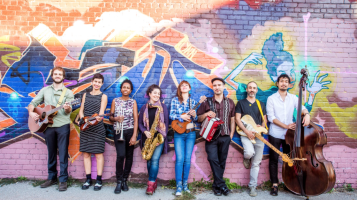 Six members of The Blue Dahlia stand in frnt of a mural depicting mountains and sky in lavender, orange, yellow, and blue