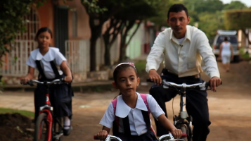 A still from The Bicycle City showing a man and two young girls wearing school uniforms riding bicycles along a dirt road.