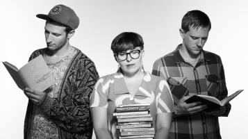 The three members of the Leatherbound Books, seen in black and white, stand together, as two of them read books and one holds a large stack of books.