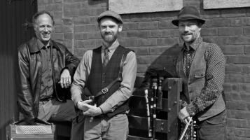 The three members of Triton stand in front of a brick wall, one holds a fiddle, and one holds an accordian.
