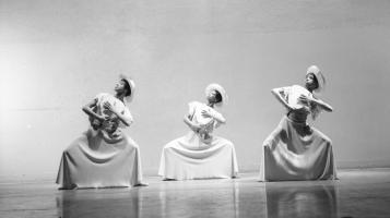 Three dancers from the Alvin Ailey company plié while holding fans and wearing long dresses and sunhats.