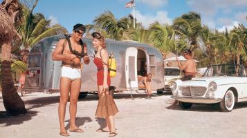 A man in white swim trunks and a woman in a red bathing suit stand in front of an airstream trailer.