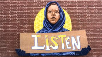 A mural depicting a young black boy wearing a navy blue hoodie, holding a sign that says "Listen" with the sun shining behind him.