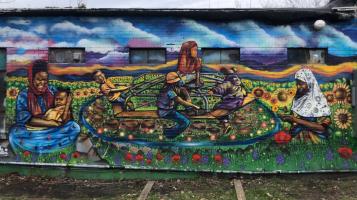 A colorful mural painted on a brick wall depicting a black woman wearing a hijab and holding a baby, children on a spinning playground structure, and a black teenager wearing a white hijab, all centered in a field of sunflowers.