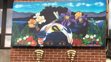 A mural that shows a black woman's face from the eyes up, with tropical flowers in her hair, and mountains and a lake in the background. Large gold earrings hang below the mural. 