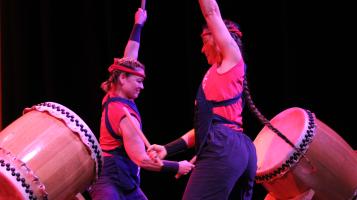 Two women with their arms raised, ready to strike two large taiko drums