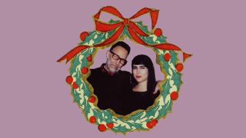 A photograph of Kat Wright and Brett Hughes from the shoulders up encircled with an illustrated wreath with red berries and a red bow on a lavender background