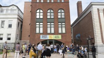 People stand in small groups talking to each other on the back patio of The BCA Center, a narrow 4 story, red brick building with tall, arched windows.