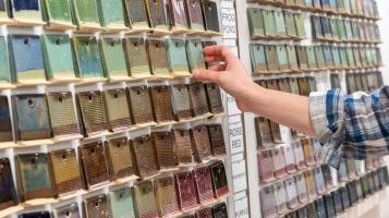 The hand of a light skinned person wearing a plaid shirt reaches toward a wall of clay tiles with various colors of glazes