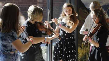 Five young women stand together, three playing the fiddle, and one playing the flute