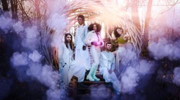 A photo of a group of people wearing white, futuristic clothes, standing in an orb woven from tree branches surrounded by purple clouds