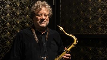 Alex standing with saxophone in front of vintage wall paper