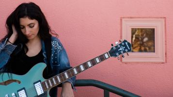 Ali T holding a vintage teal guitar in front of a pink concrete wall
