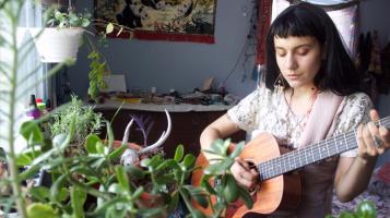 Hana is playing an acoustic guitar, with her eyes closed, surrounded by house plants