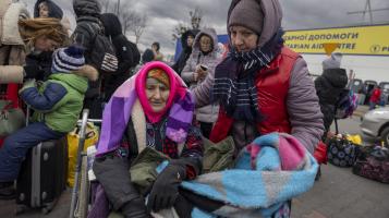 A photograph of an elderly woman sitting in a wheelchair, bundled in many multicolored layers of scarves and coats, being attended to by a woman wearing a grey parka and a red vest, with other people and a yellow and blue sign with Cyrillic writing in the background