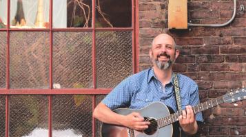 Phil holding an acoustic guitar in front of brick wall with a beautiful window