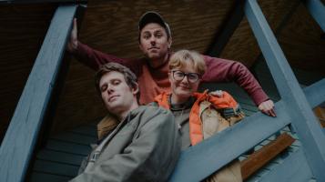 Two light skinned men and one light skinned woman with short blonde hair and glasses stand close together on an outdoor staircase with blue handrails, as seen from below.