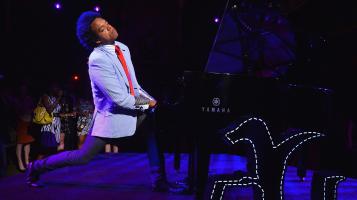 Eric Lewis, a dark skinned man wearing a dove grey blazer, stands with his head thrown back, playing a black grand piano