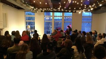 A crowd of people, seen from behind sit in a room watching a person standing at a lectern. Twinkle lights are strung on the ceiling.