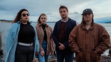 Three women and one man stand on the shore of a lake. One woman wears round sunglasses and a light blue coat over a black top, one wears a fawn colored jacket and one wears a brown jacket and a baseball cap. The man wears a dark blue blazer over a burgundy sweater