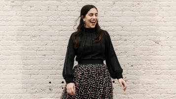 A light skinned woman wearing a black high necked blouse with full sleeves, and a black shirt with small multi-colored polka dots laughs and stands in front of a brick wall painted white