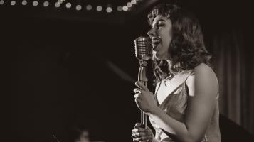 A black and white photograph of a light skinned woman with shoulder length dark, wavy hair singing into a microphone