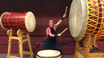 A light skinned man wearing a black bib vest over a red tie dyed shirt, with a red headband holds two large drum sticks and stands ready to strike the oversized drums on either side and in front of him.