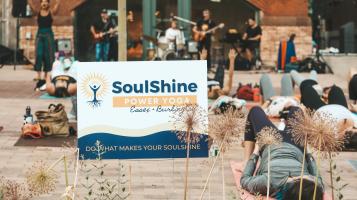 A sign that says Soulshine Yoga in the foreground with people laying down on yoga mats on a brick surface and band in the background