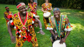 In the foreground, two dark skinned men stand on a green lawn, wearing colorful traditional African clothes, while one play a drum and one dances. In the background two dark skinned women and one light skinned women wear colorful traditional African clothing and play drums