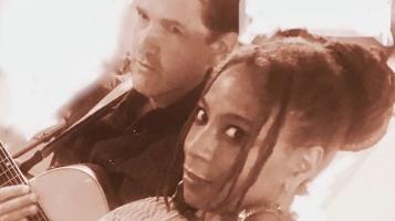 A brownish red tinted image of a light skinned man holding a guitar, and a dark skinned woman, tilted at an angle with a white background