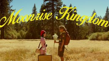Moonrise Kingdom, written in sweeping yellow text arching over an image of a boy wearing a coonskin cap holding flowers facing a girl wearing a red hat and holding a brown suitcase. They stand in the middle of a field of gold grass, with evergreen trees and blue sky in the background.