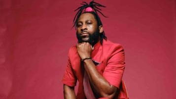 A dark skinned man wearing a red blazer, and dreadlocks in a top knot secured with a red band, sits in front of a red background, resting his chin on his hand