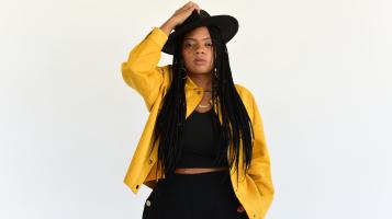 A dark skinned woman with long cornrow braids, wears a yellow jacket, black crop top and pants, and holds her hand up to her black wide brimmed hat