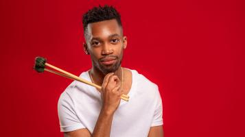 A dark skinned man sits in front of a red background, wearing a white tee shirt hold vibraphone sticks