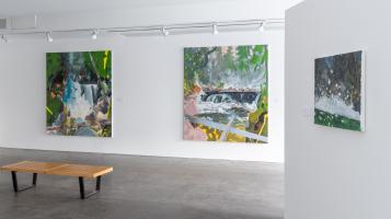 Two large and one smaller paintings of abstract forest scenes hang on white gallery walls, with a wooden bench in the foreground
