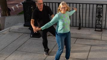 A man wearing a black shirt and a woman wearing an aqua sweater hold hands and dance outdoors in City Hall Park