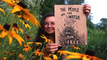 a photograph of a woman with long brown hair and glasses sitting in a field of black eyed susans holding a print that says The People and the water will flow free with illustrations