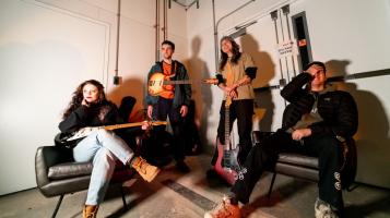 A light skinned woman with wavy brown hair and three light skinned men sit together in an industrial space with white walls