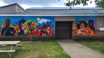 A mural mounted on a low, cement building that depicts dark skinned adolescent boys and girls, outlined in vibrant colors against a bright blue sky background with large, stylized orange, pink and red flowers