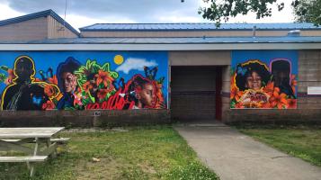 A mural mounted on a low, cement building that depicts dark skinned adolescent boys and girls, outlined in vibrant colors against a bright blue sky background with large, stylized orange, pink and red flowers 