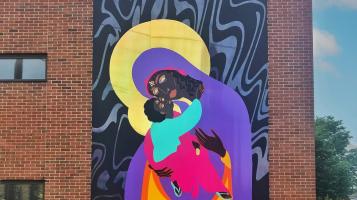 A mural mounted on a brick building depicts a dark skinned madonna and child, with the mother wearing a purple, pink and yellow robe and the child held in her arms, reaching for her bowed head wearing an aqua tunic. The background is black and grey marbleized swirls