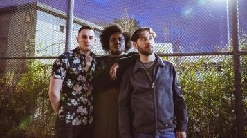 Two light skinned men and a dark skinned woman stand close together in front of a tall chain link fence at twilight, with green grass and a darkening sky behind