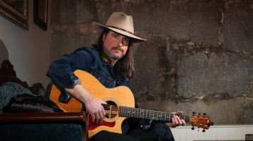 A light skinned man wearing a beige cowboy hat sits holding a honey colored acoustic guitar in front of a cracked concrete wall