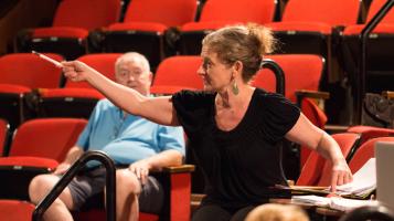 A light skinned woman with medium blond wavy hair pulled back in a bun sits a theater with red seats and gestures with a pencil in her hand