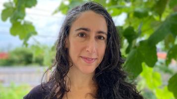 A headshot of a light skinned woman with wavy dark hair lightly streaked with greay, with green leaves in the background.